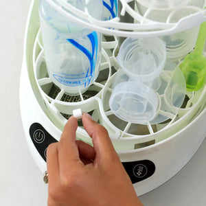 A close-up of a breastmilk bottle and pump sterilizer showing a hand dropping a tablet into the open compartment containing clear silicone bottles and pump parts.