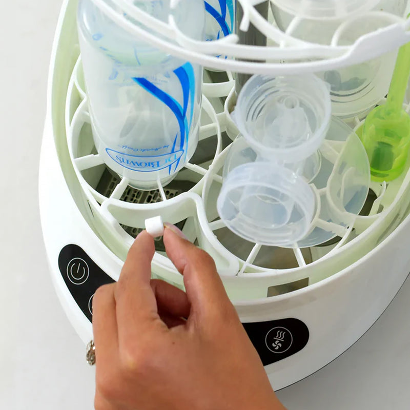 A close-up of a breastmilk bottle and pump sterilizer showing a hand dropping a tablet into the open compartment containing clear silicone bottles and pump parts.
