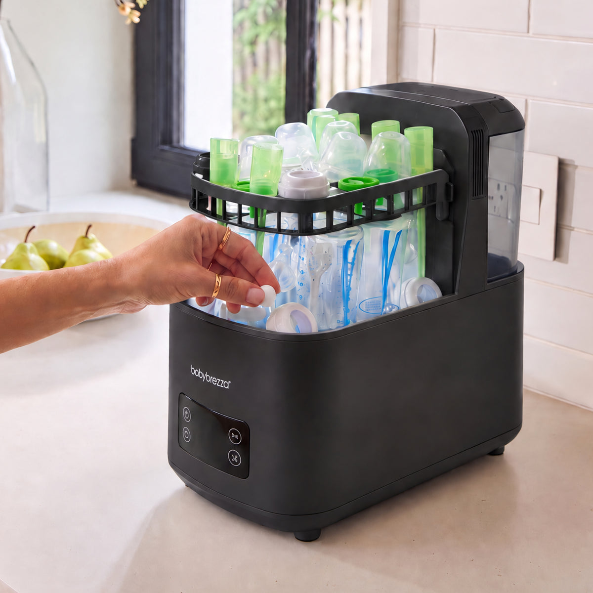 A countertop baby bottle cleaning and sterilizer machine in matte black, shown with a hand placing baby bottle detergent tablets into the interior. The machine is set on a light beige kitchen counter with a tiled wall and a window in the background.
