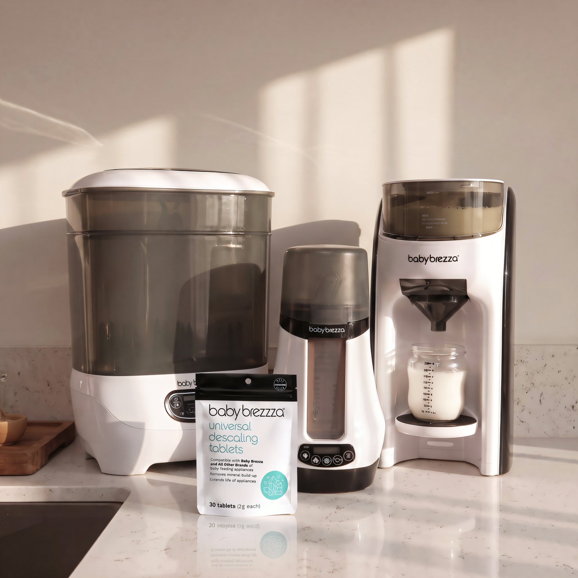 Three Baby Brezza baby care devices and a box of universal descaling tablets arranged on a white countertop in a kitchen setting with warm sunlight casting shadows.