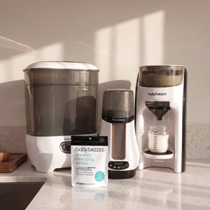 Three Baby Brezza baby care devices and a box of universal descaling tablets arranged on a white countertop in a kitchen setting with warm sunlight casting shadows.