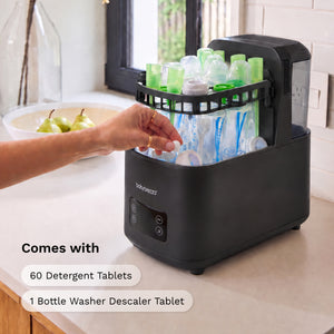 A countertop bottle washer and dryer combo in matte black, shown with a hand placing detergent tablets into the top rack. The unit is set in a bright kitchen environment, with text overlays stating 'Comes with 60 Detergent Tablets 1 Bottle Washer Descaler Tablet'.
