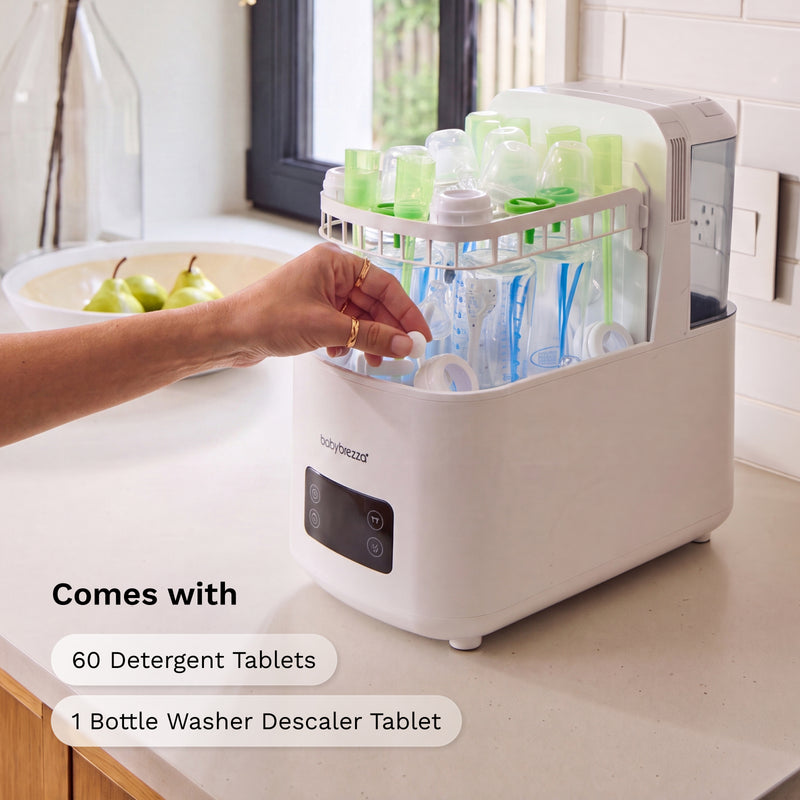 A white bottle washer and dryer combo machine on a kitchen counter, with a hand reaching toward the front control panel. The top rack holds baby bottles and cups, and text on the image reads 'Comes with 60 Detergent Tablets 1 Bottle Washer Descaler Tablet'.