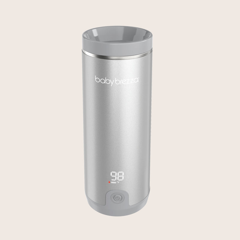 A silver Baby Brezza travel warmer with a digital temperature display showing 98 degrees and a power button, set against a solid light beige background.