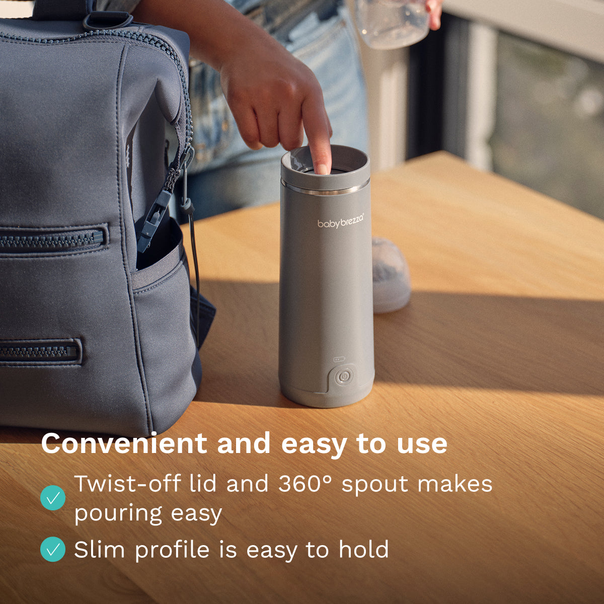 Gray Baby Brezza Portable Warmer with a twist-off lid on a wooden surface, next to a backpack.
