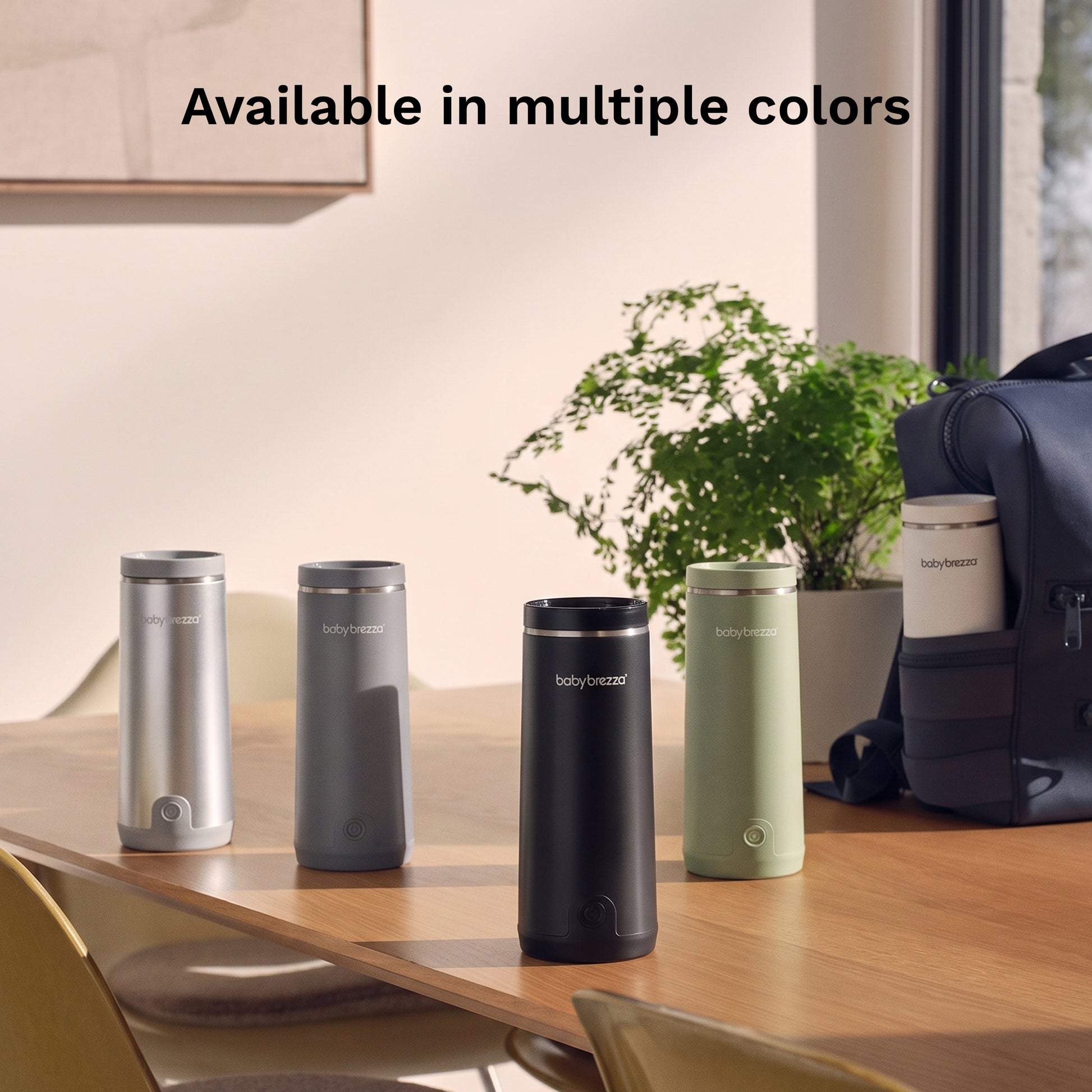 Five Baby Brezza Portable Warmers in different colors (silver, dark gray, black, white, and sage green) lined up on a wooden table in a modern room setting, with a plant and a backpack in the background. Text at the top reads 'Available in multiple colors'.