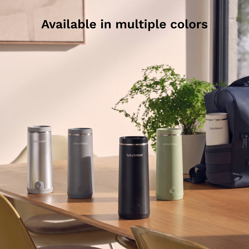 Five Baby Brezza Portable Warmers in different colors (silver, dark gray, black, white, and sage green) lined up on a wooden table in a modern room setting, with a plant and a backpack in the background. Text at the top reads 'Available in multiple colors'.