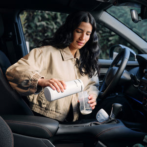 Woman inside a car holding a white Baby Brezza Portable warmer and a clear baby bottle.