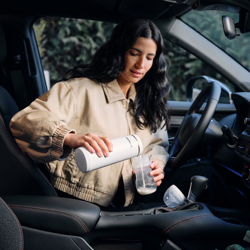 Woman inside a car holding a white Baby Brezza Portable warmer and a clear baby bottle.