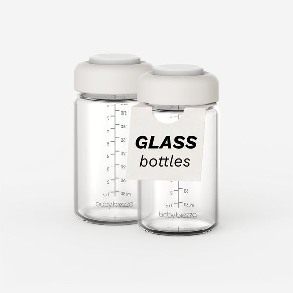 Two clear replacement glass baby bottles with white caps, featuring black measurement markings and the brand name 'baby brezza' near the base, with a white label reading 'GLASS bottles' in the foreground.